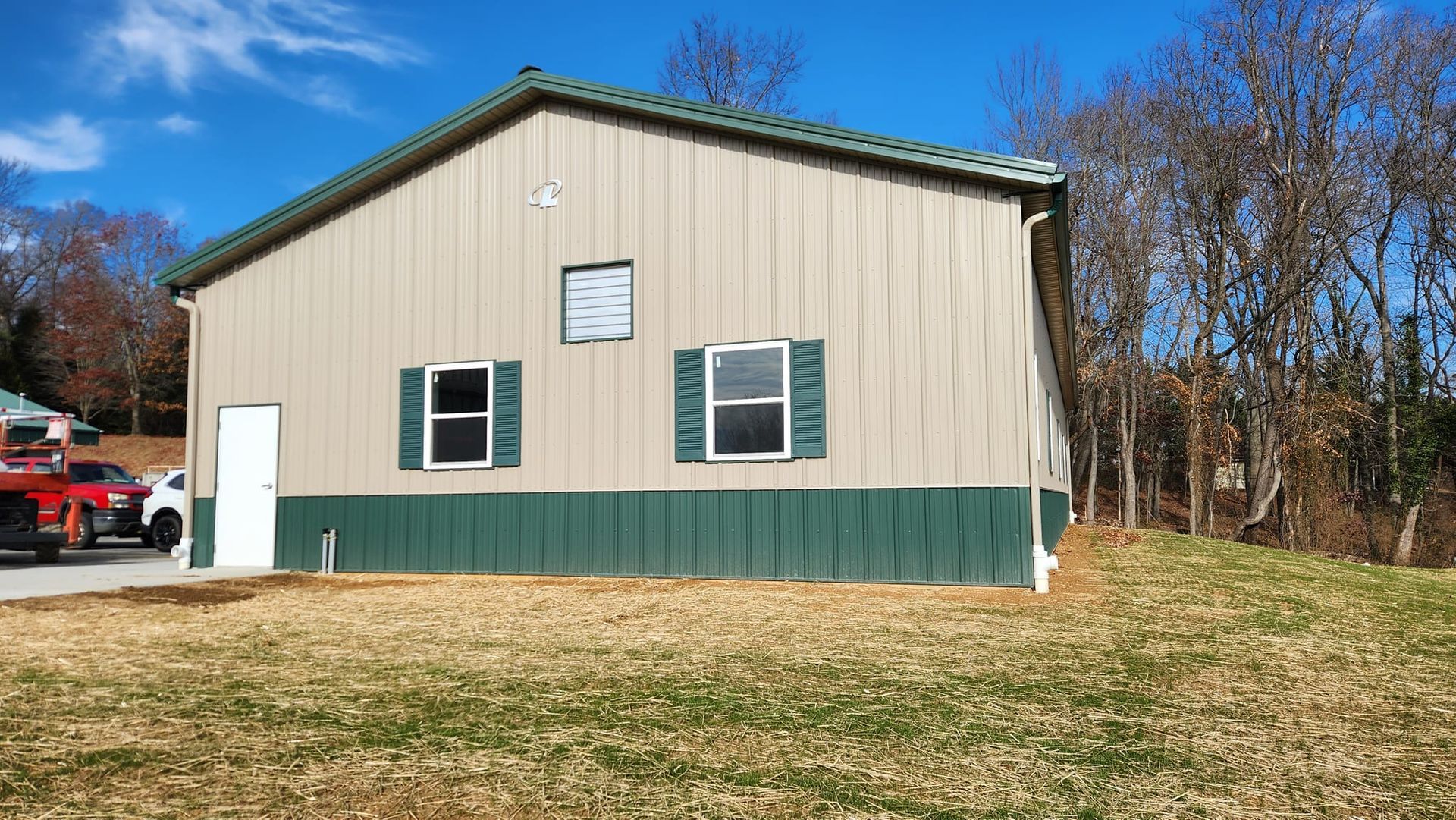 A tan metal building with green trim, dark green shutters, and a white door, set in a grassy field against a blue sky.