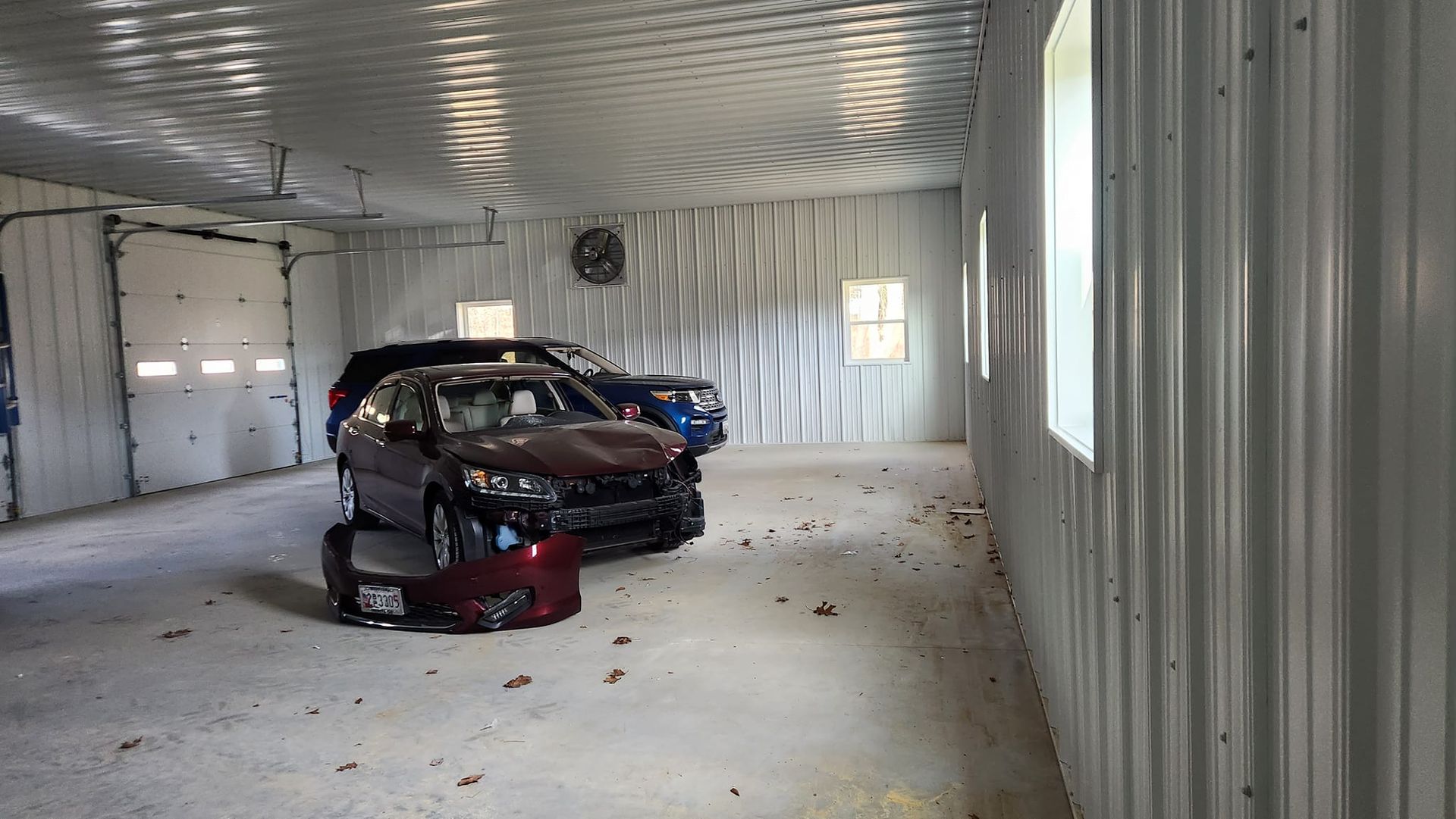 A maroon car with front-end damage and a detached bumper parked inside a large, metal-walled garage with concrete floors.