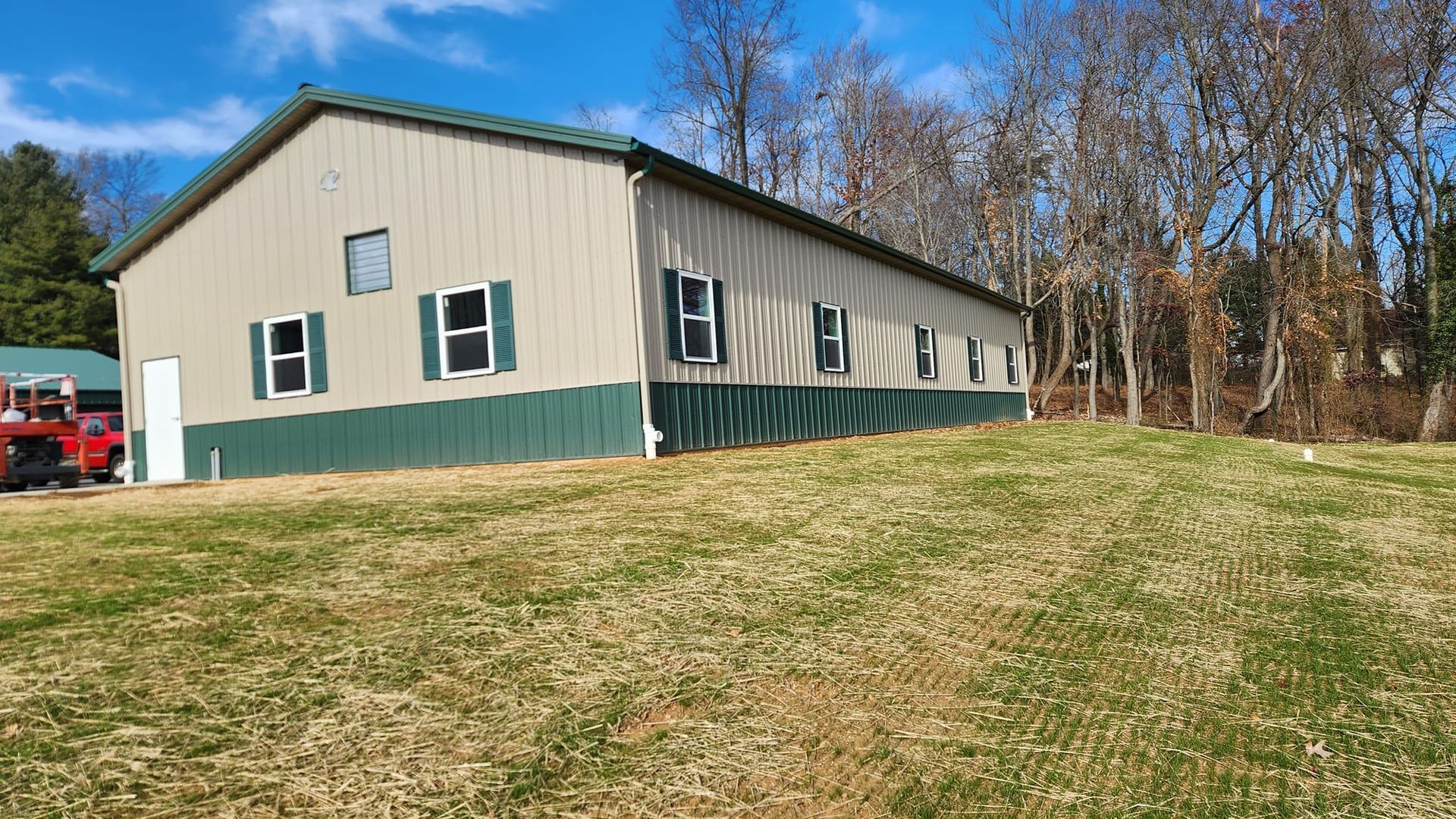 A tan and green metal building sits on a grassy hill under a blue sky, with trees visible in the background.