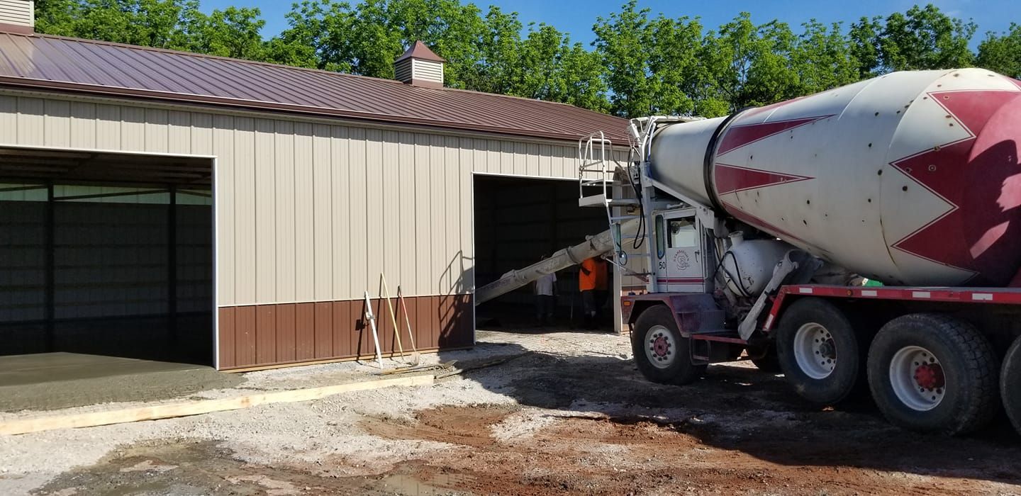 A cement mixer truck pours concrete into the open doorway of a tan and brown metal building on a sunny day.