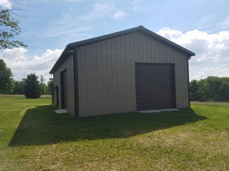 A tan, metal-sided storage building with a large brown garage door and a side entry door, situated in a grassy field.