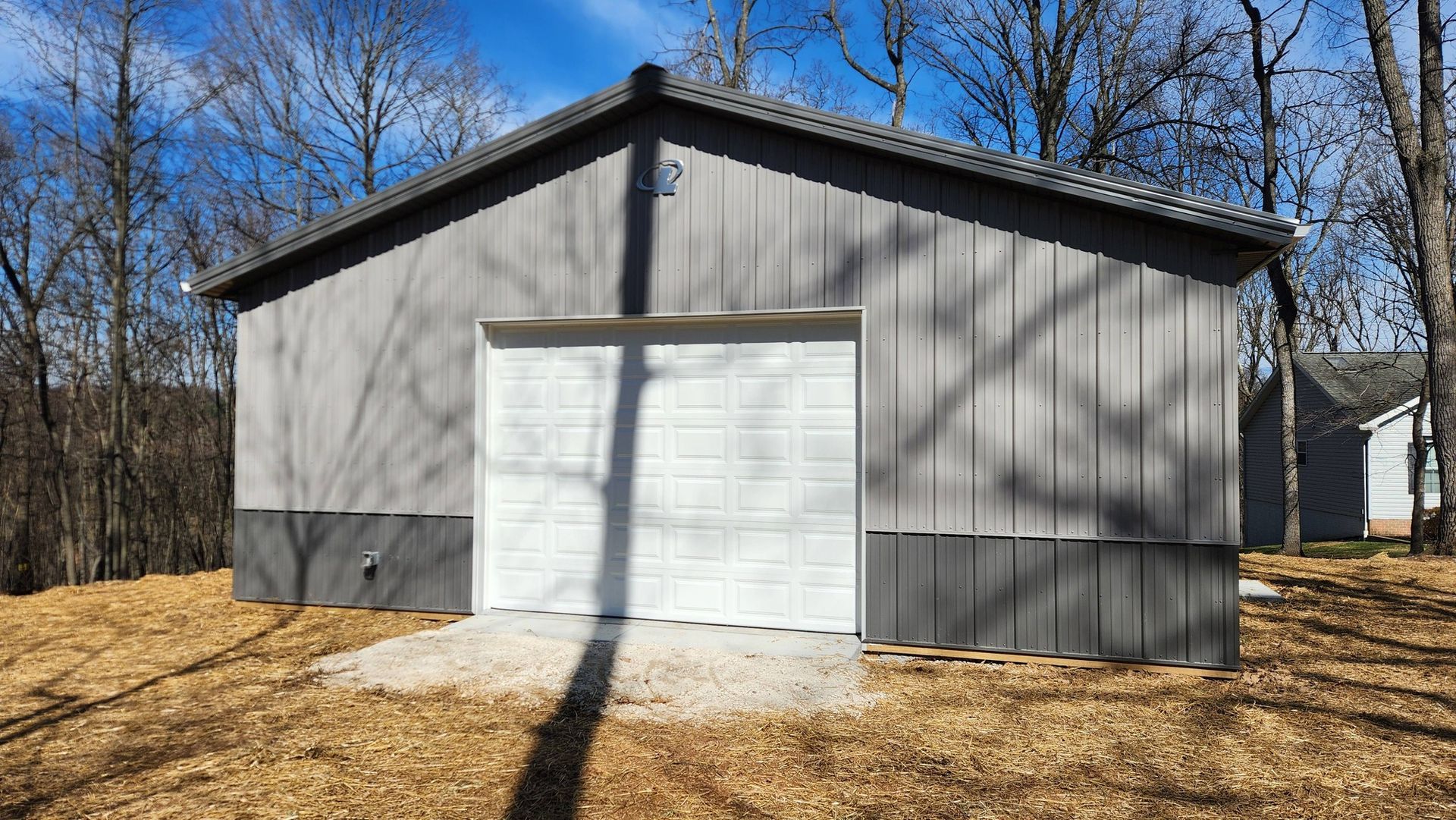 A gray, gabled workshop with a white garage door, nestled in a wooded area with dry grass under a bright blue sky.