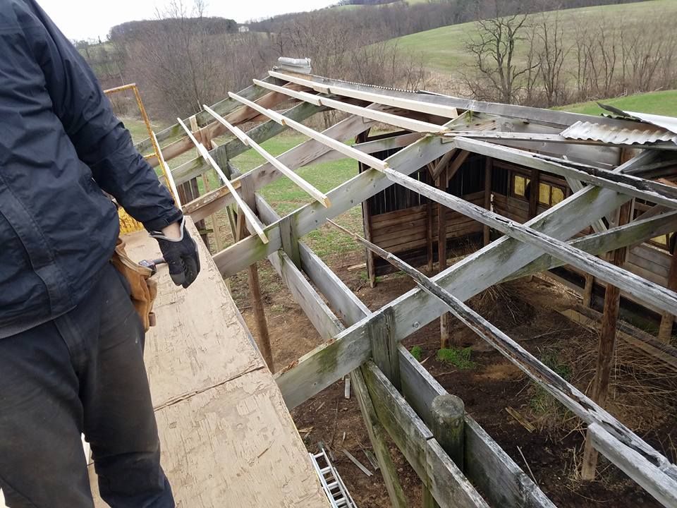 A person in a dark jacket stands on scaffolding, working on the wooden roof rafters of an outdoor shed.
