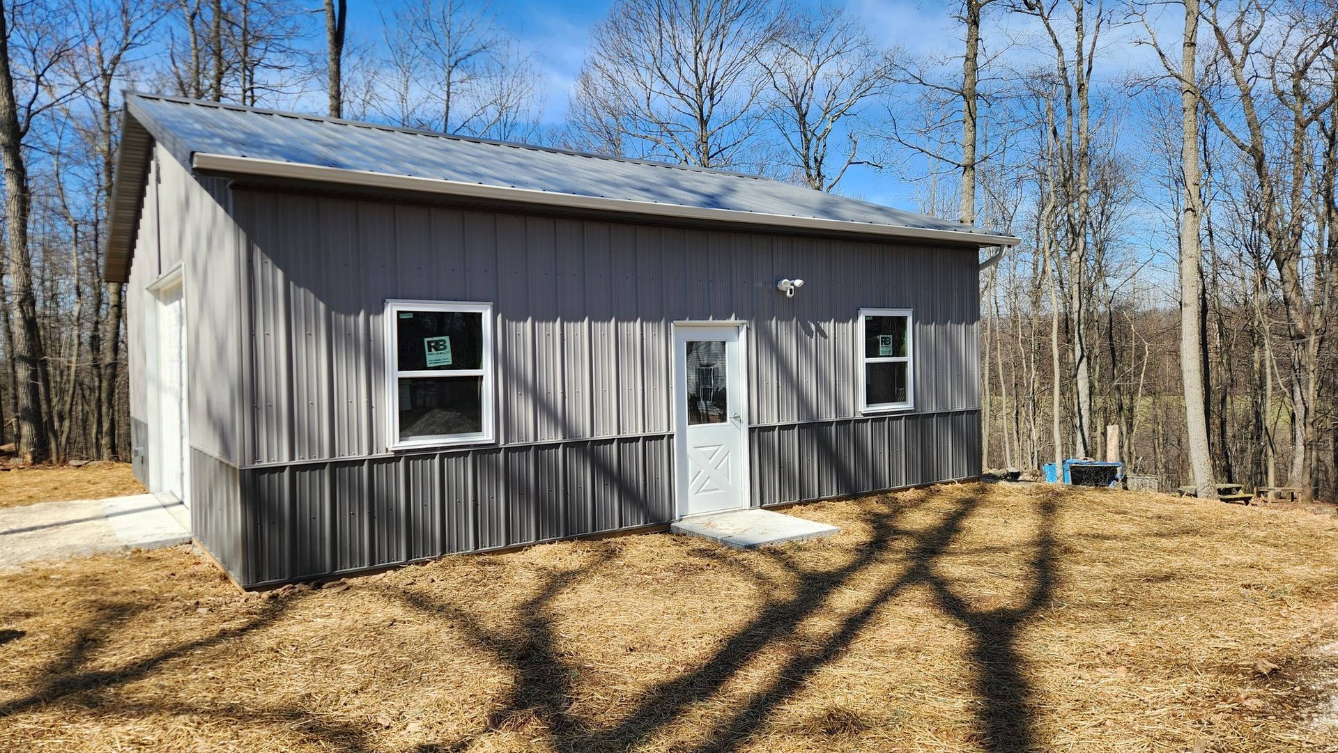 A rectangular metal building with light grey siding and a dark grey lower panel, set among bare trees on a sunny day.