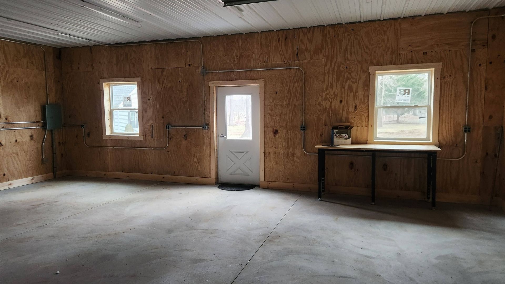 An interior view of a garage with plywood walls, a center door, two windows, and a workbench on the right.