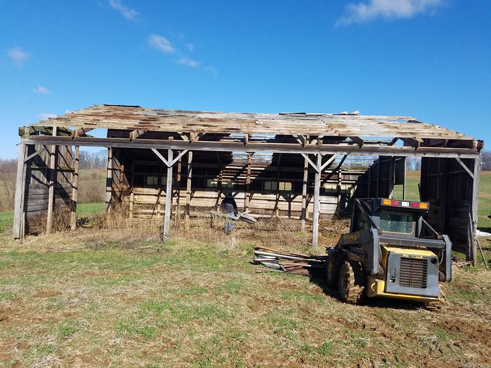A yellow skid steer sits on grass in front of an old, partially collapsed wooden barn under a blue sky.