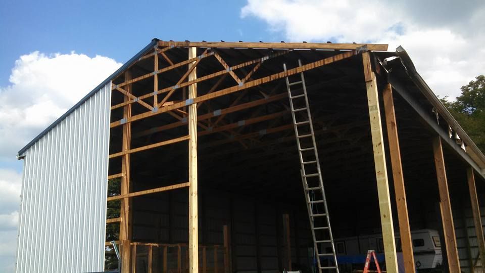 A wooden-framed pole barn under construction with partially installed white metal siding and a ladder leaning inside.