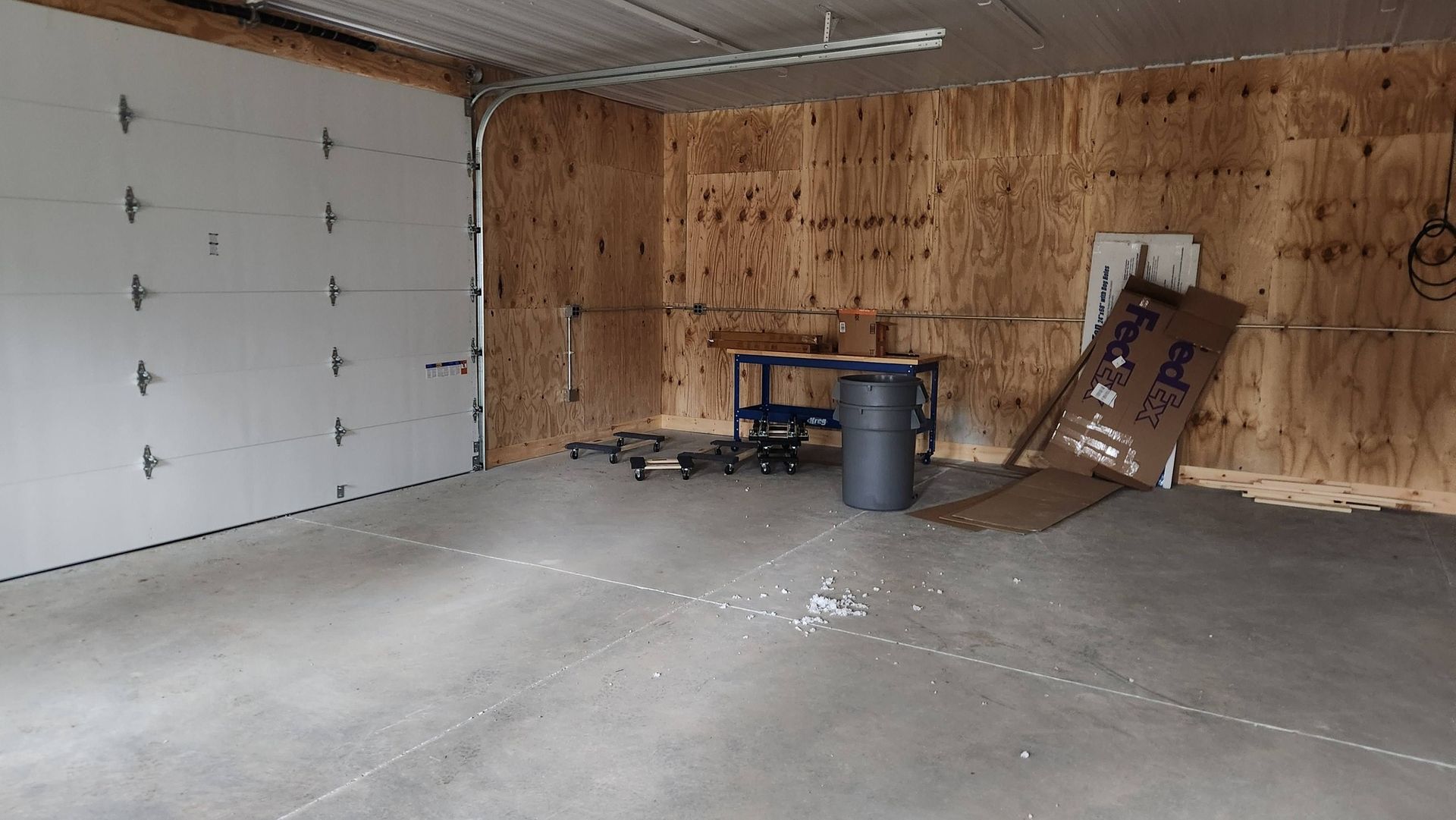 An empty, unfinished garage with plywood walls, a white overhead door, a concrete floor, and a metal work table.