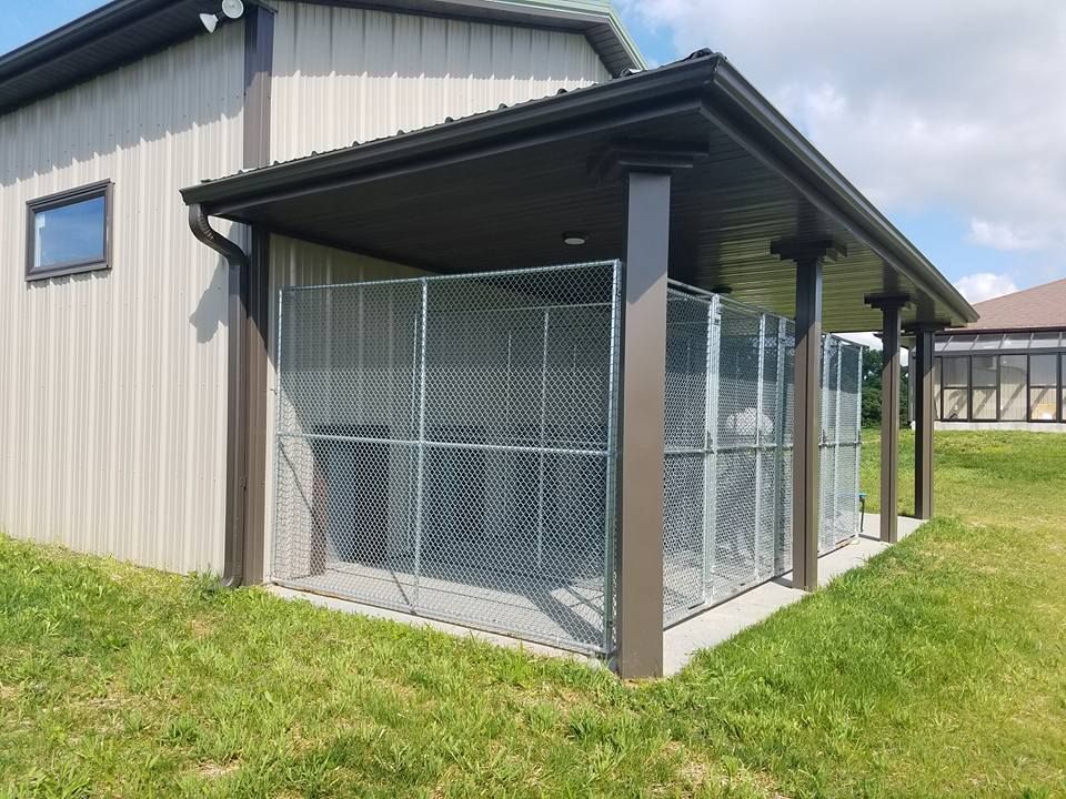 A grey metal building with a covered patio area containing a large, wire-mesh kennel enclosure on a concrete pad.