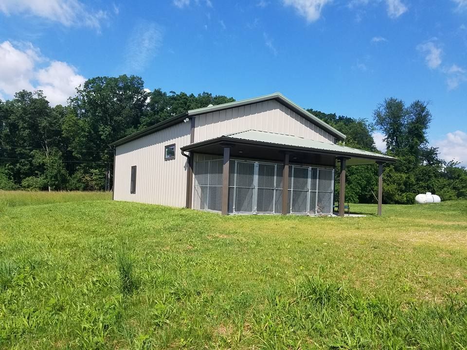 A beige metal outbuilding with a screened-in porch, set in a grassy field under a sunny blue sky with trees.
