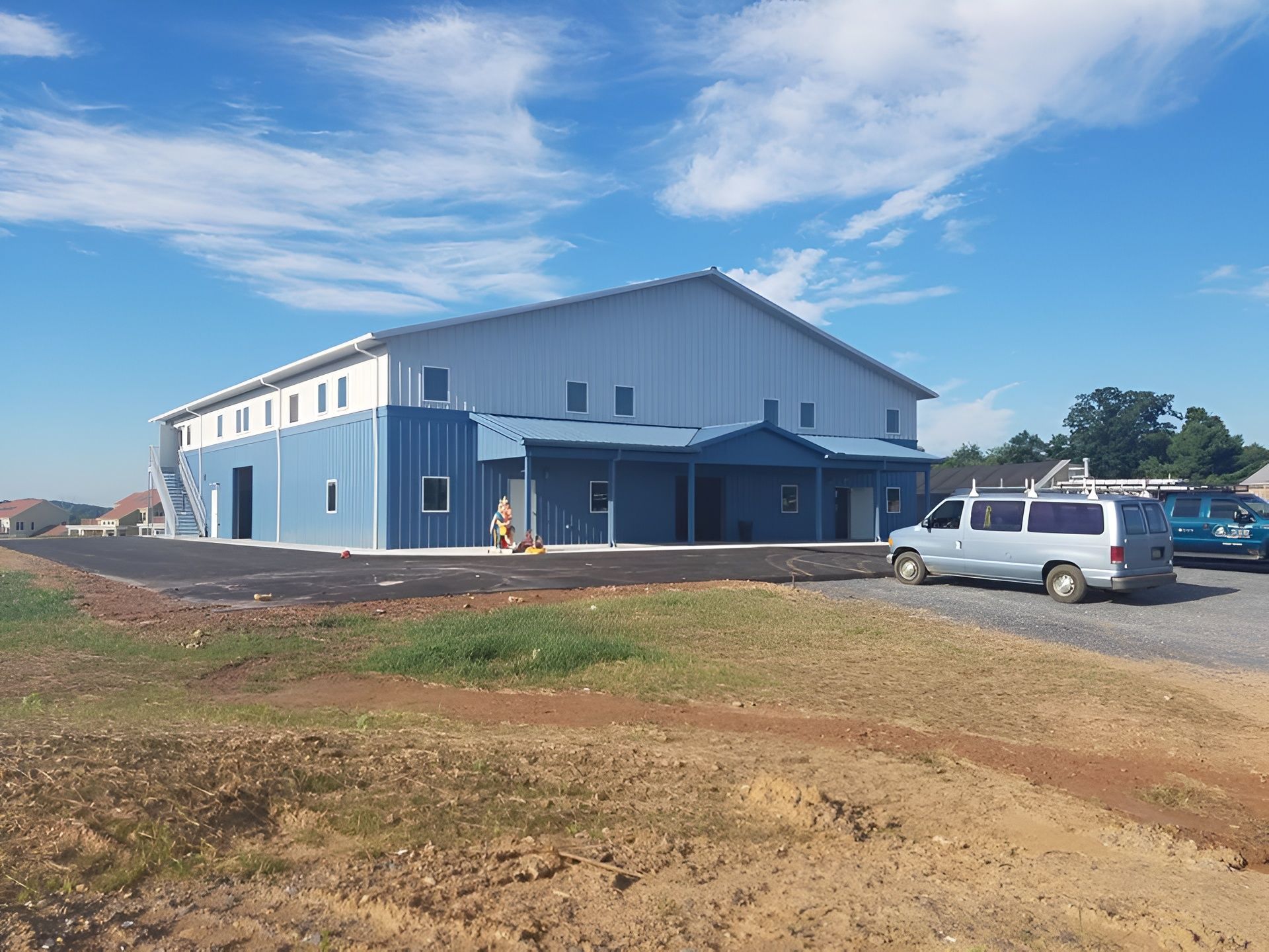 A two-story blue and grey metal building with an attached porch, next to a parking area under a blue sky.