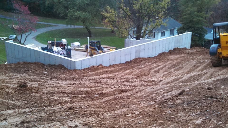 Construction site featuring a new concrete retaining wall on a sloped, muddy lot near a house and trees.