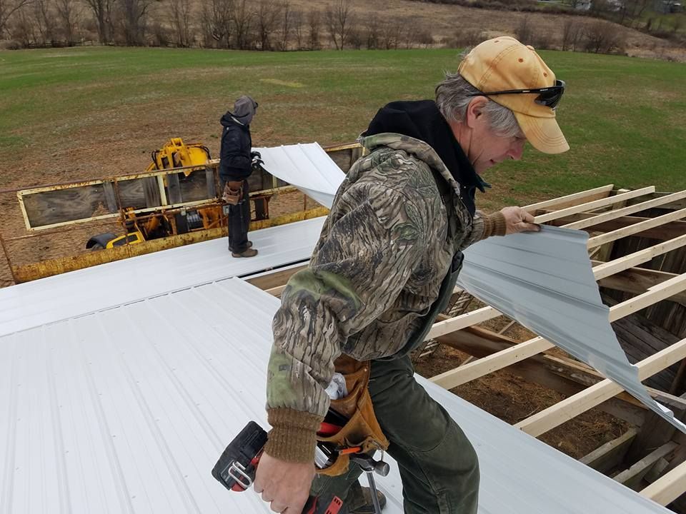 Two people in outdoor clothing install white metal roofing panels onto the wooden frame of a building in a grassy field.