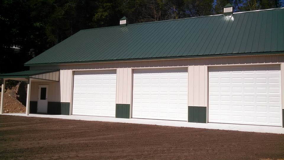 A light-colored pole barn with a dark green metal roof, three white garage doors, and a small porch on the left.