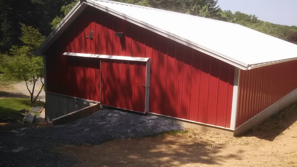A red metal-sided barn with a white roof sits on a sloped, gravel-covered lot near trees.