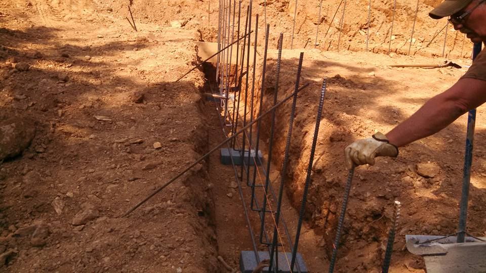 A construction worker uses a tool to position vertical rebar stakes in a trench dug into dry, earthen ground.