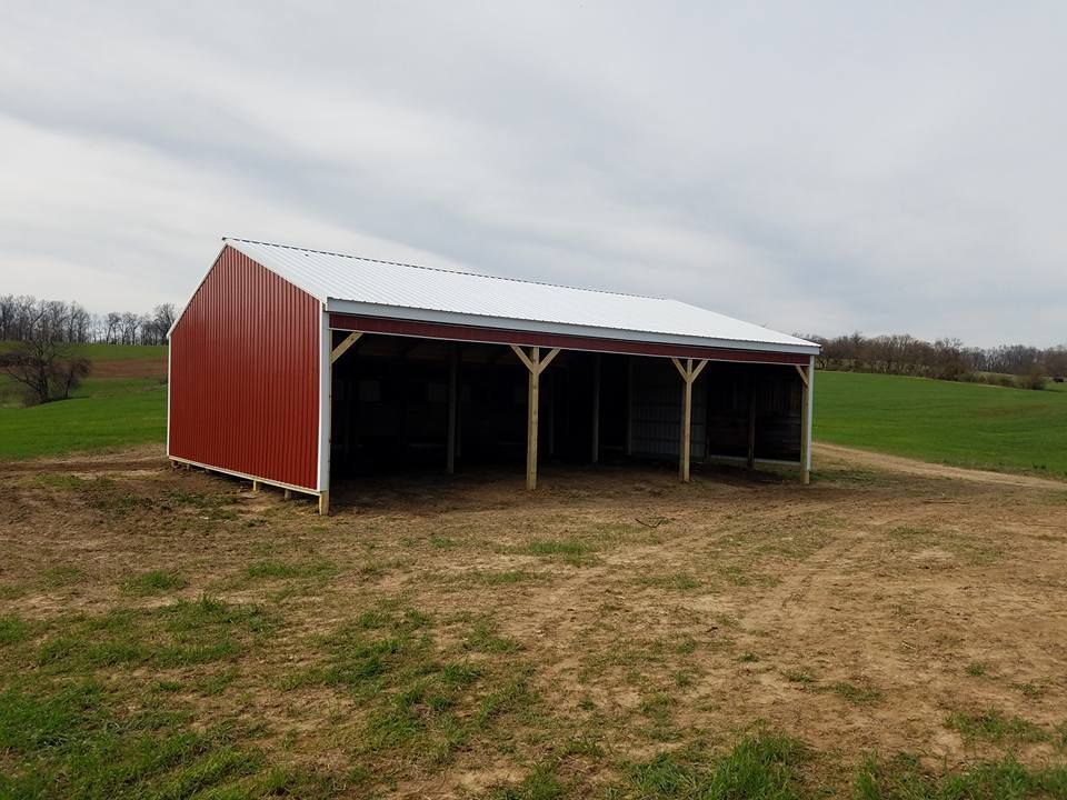 A red-sided, open-front pole barn with a white roof stands in a grassy field under a cloudy sky.
