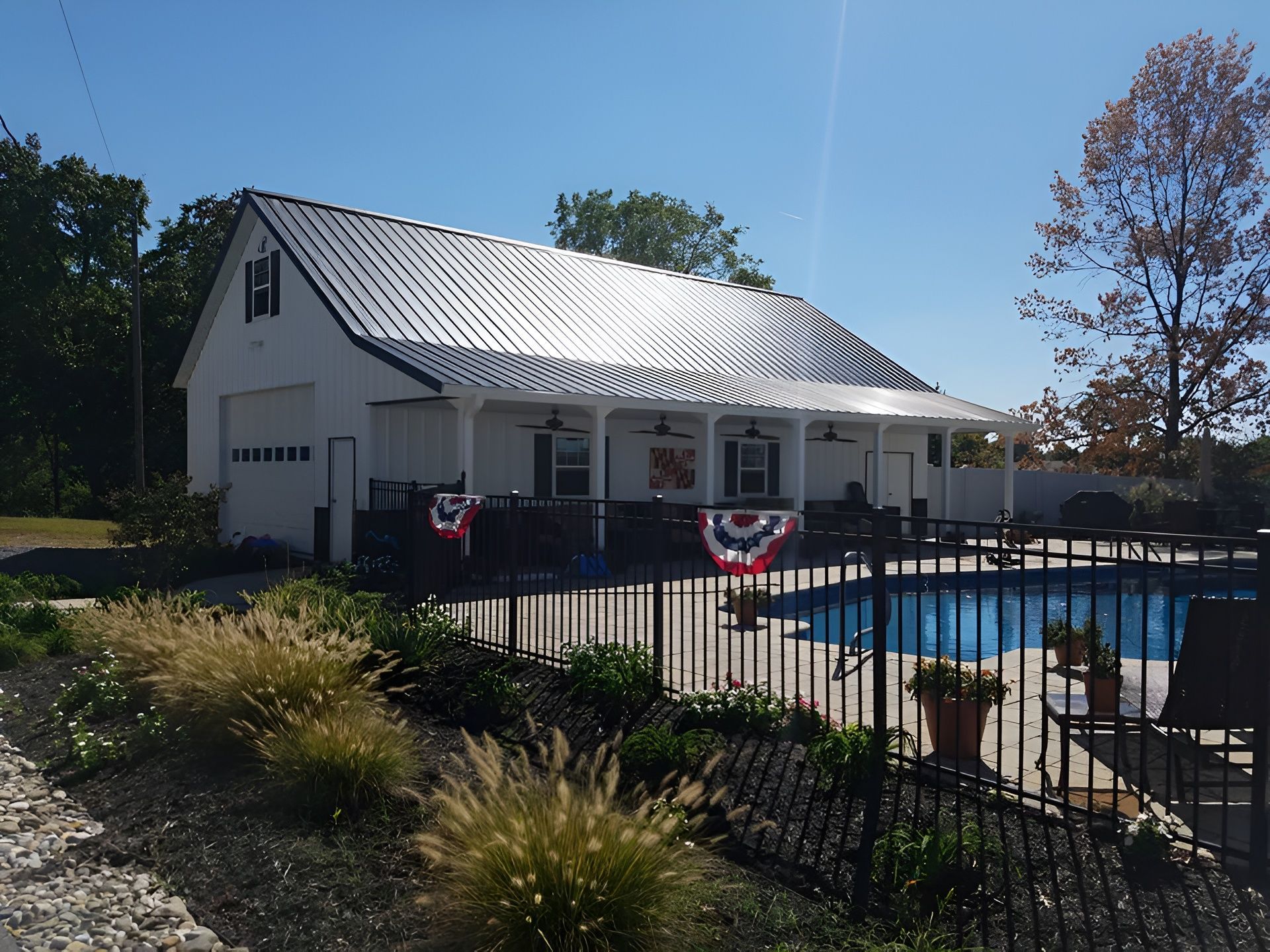 A white detached garage with a dark metal roof and a covered patio overlooking a swimming pool on a sunny day.