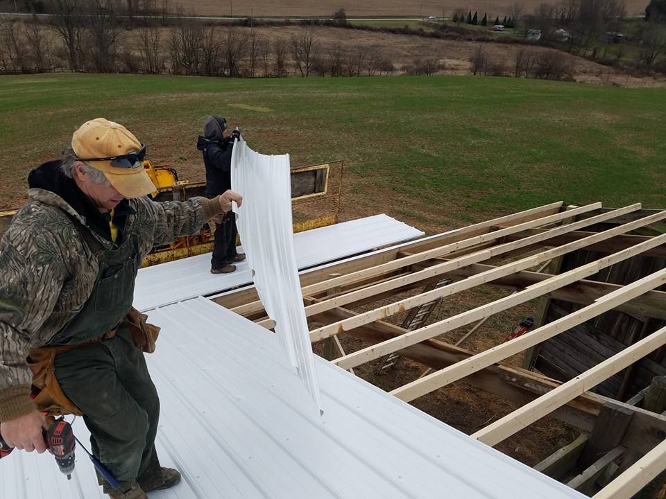 Two people in work clothes install white metal roofing panels on the wood frame of a building in a rural, grassy field.