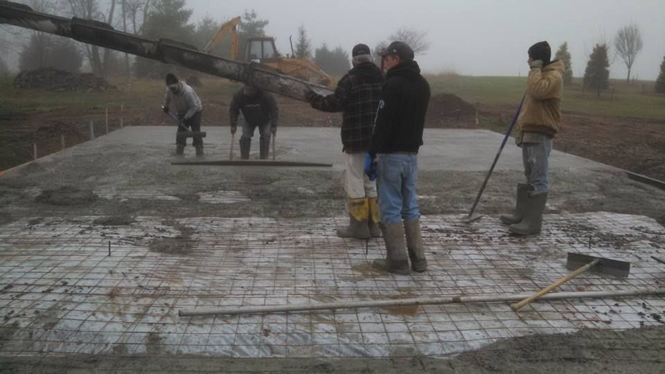 Five people in work clothes stand on a concrete construction site as wet cement is poured from a chute.