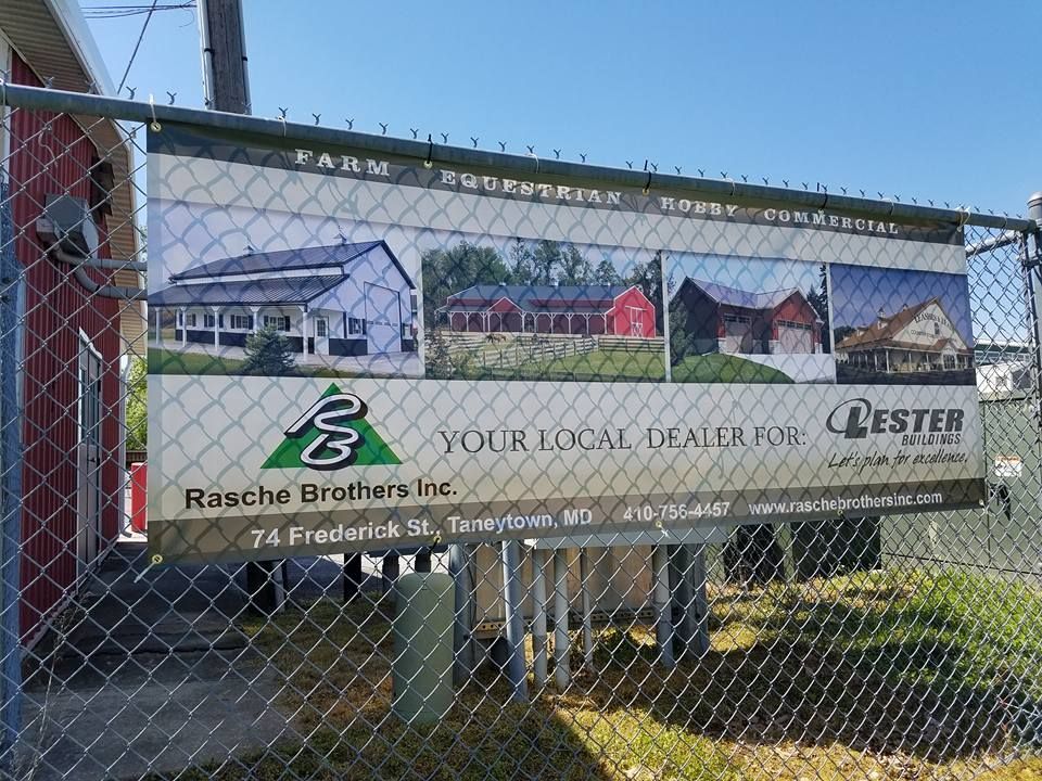 A sign on a chain-link fence for Rasche Brothers Inc., a local dealer for Lester Buildings, showcasing several structures.