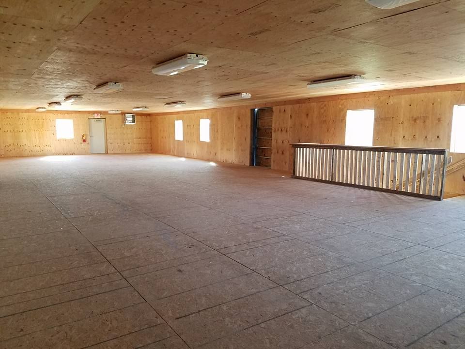 A large, empty room with light wood-paneled walls, a plywood floor, overhead lighting, and a fenced-in stairwell.