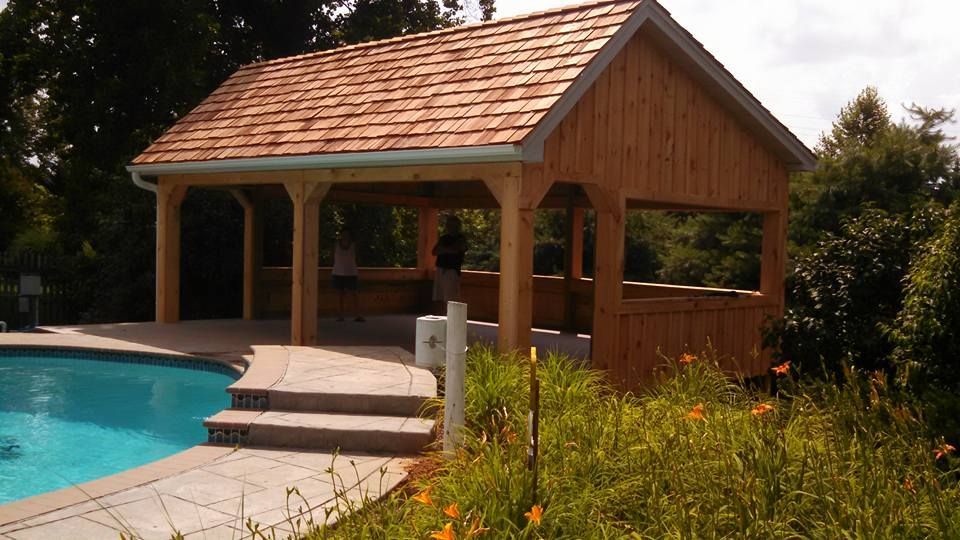 A wooden pavilion with a shingled roof stands next to a backyard swimming pool surrounded by a stone deck and greenery.