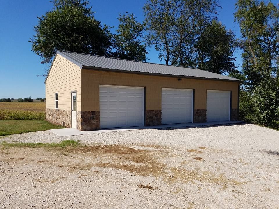 A tan three-car garage with stone siding and a metal roof stands on a gravel lot near a field and trees.