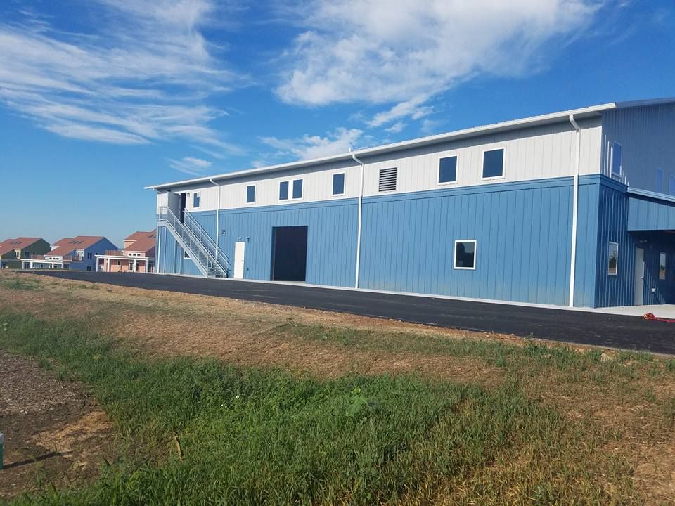 A large metal warehouse building with blue and white walls sits beside a paved path under a clear blue sky.
