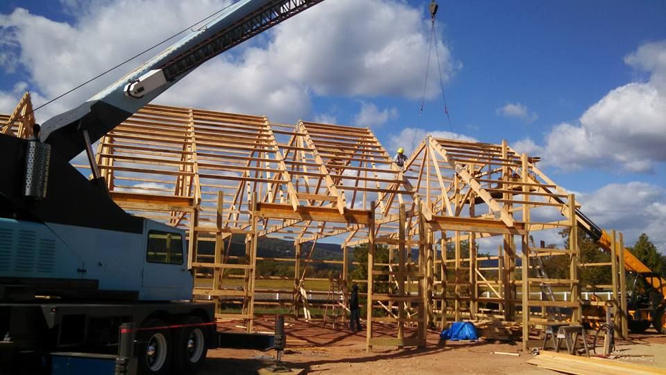 A crane lifts wooden trusses onto the timber frame structure of a building under construction on a sunny day.
