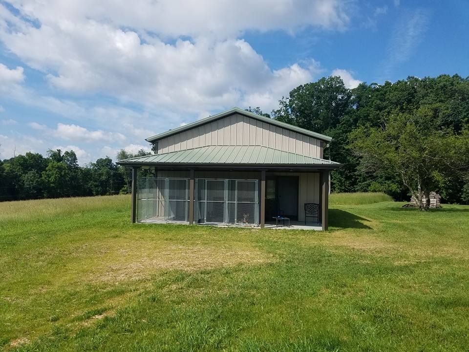 A metal-sided building with a covered front porch, glass partitions, and a green roof, situated in a grassy field.