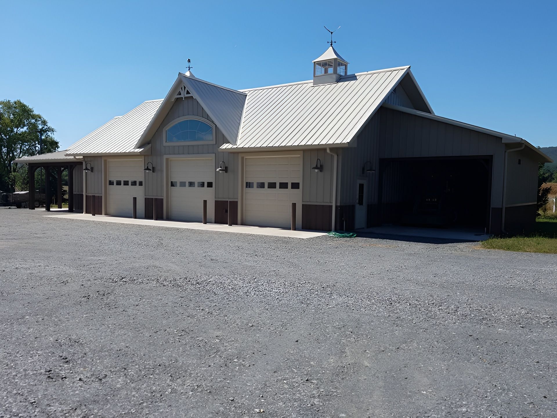 A tan building with three white garage doors and a metal roof sits beside a gravel lot under a clear blue sky.