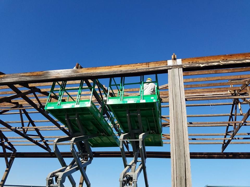 Two green scissor lifts parked side-by-side beneath the wooden rafters of an open-air structure under a blue sky.