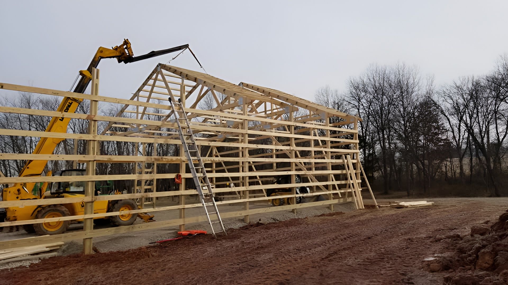 A yellow telehandler lifting a wooden truss onto the frame of a partially constructed barn in an open field.