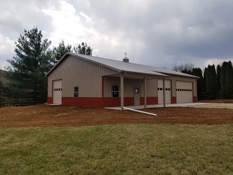 A metal-sided building with tan walls, red trim, and a porch, set on a rural property under a cloudy sky.