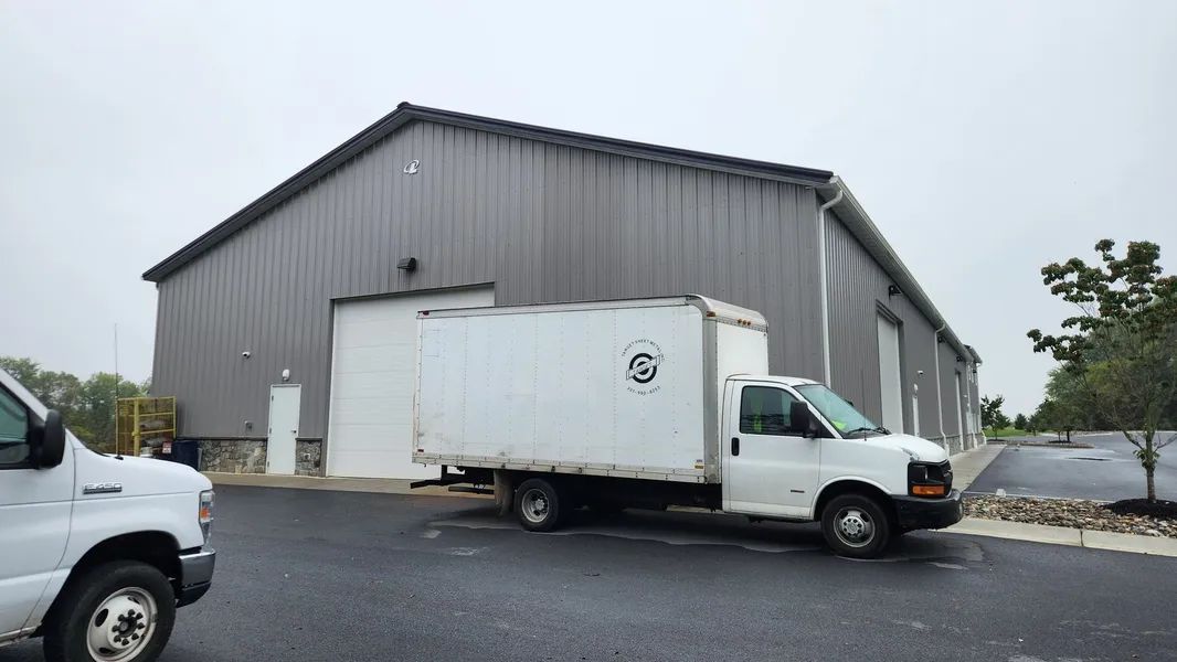 A white box truck parked in front of a gray metal warehouse building under an overcast sky.