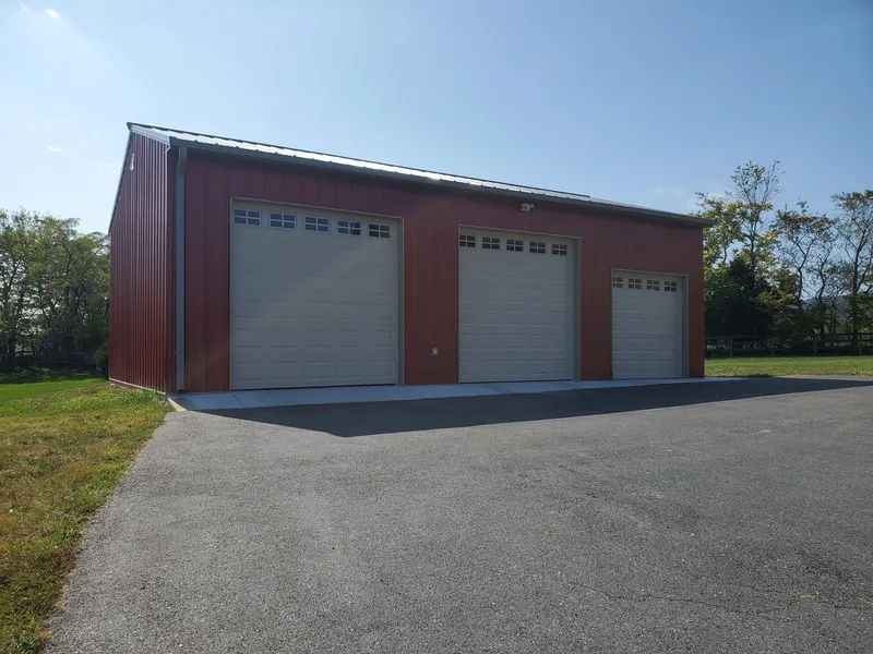 A red metal barn with three tan garage doors of varying sizes, situated on a gravel lot near trees under a blue sky.