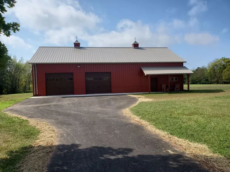 A red barn-style garage with two brown doors, a grey metal roof, and a covered side porch set on a grassy rural lot.