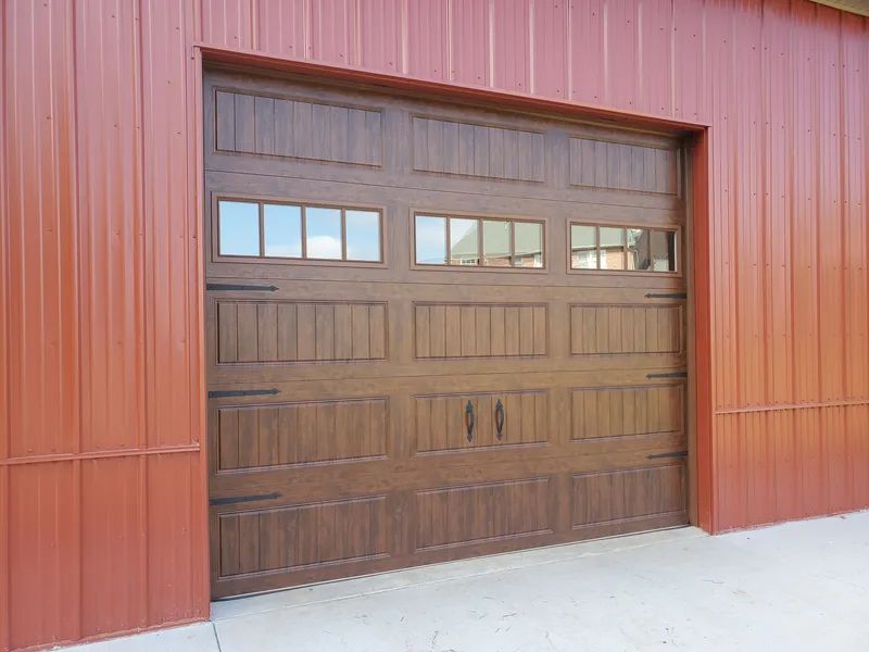 A brown carriage-style garage door with a window row, installed in a building with rust-colored vertical metal siding.