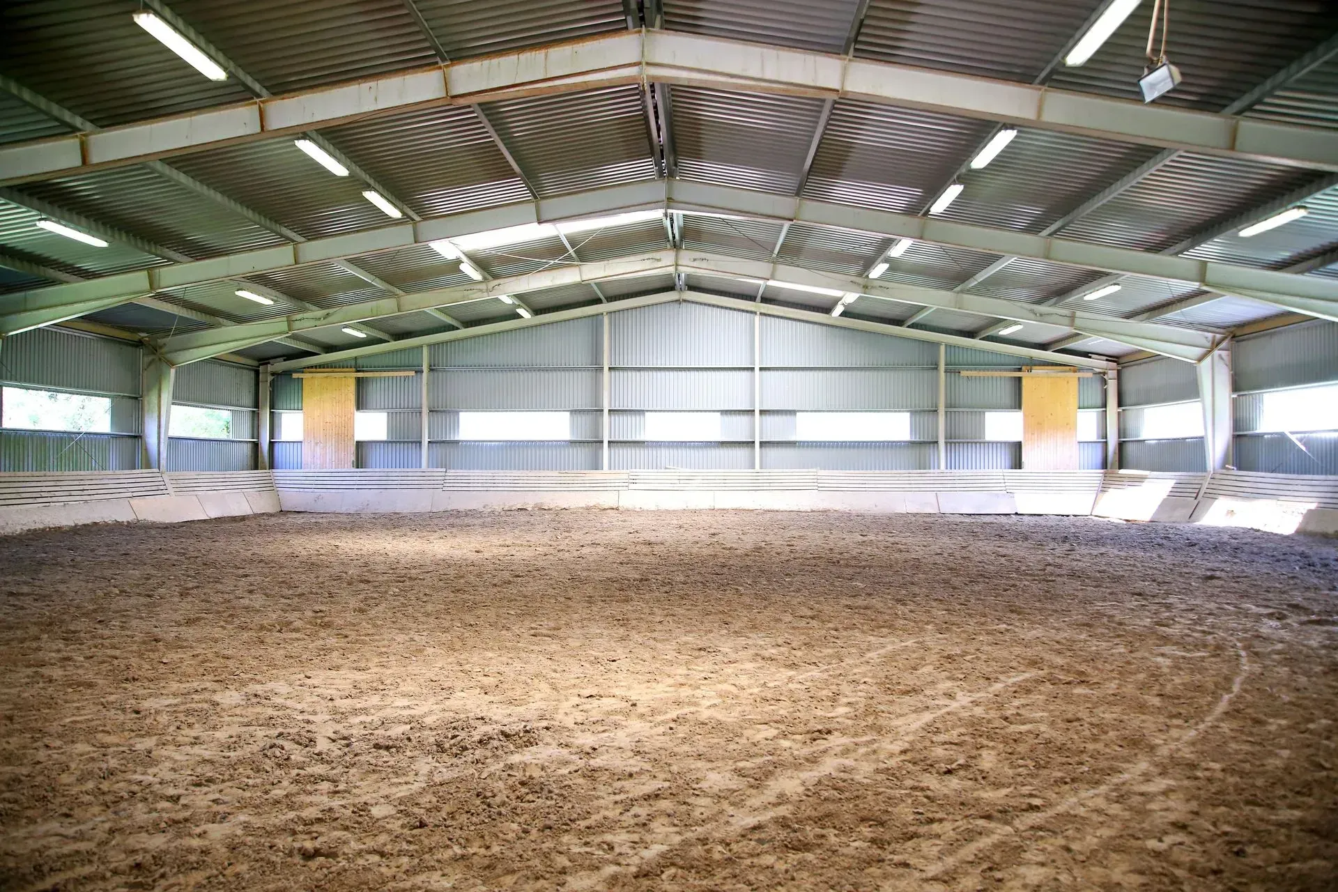 An indoor equestrian riding arena with a dirt floor, metal walls, and a high-pitched ceiling with overhead lighting.