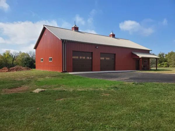 A red metal workshop with two brown garage doors and a light gray roof, situated on a grassy lot under a blue sky.