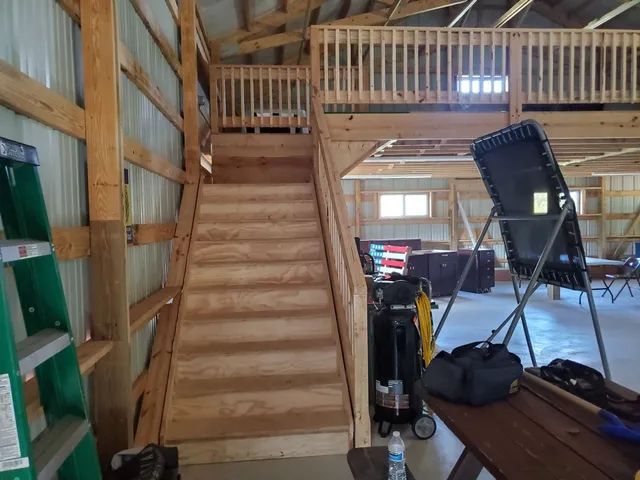 Wooden staircase leading to a loft in a workshop, with a green ladder and work equipment visible in the foreground.