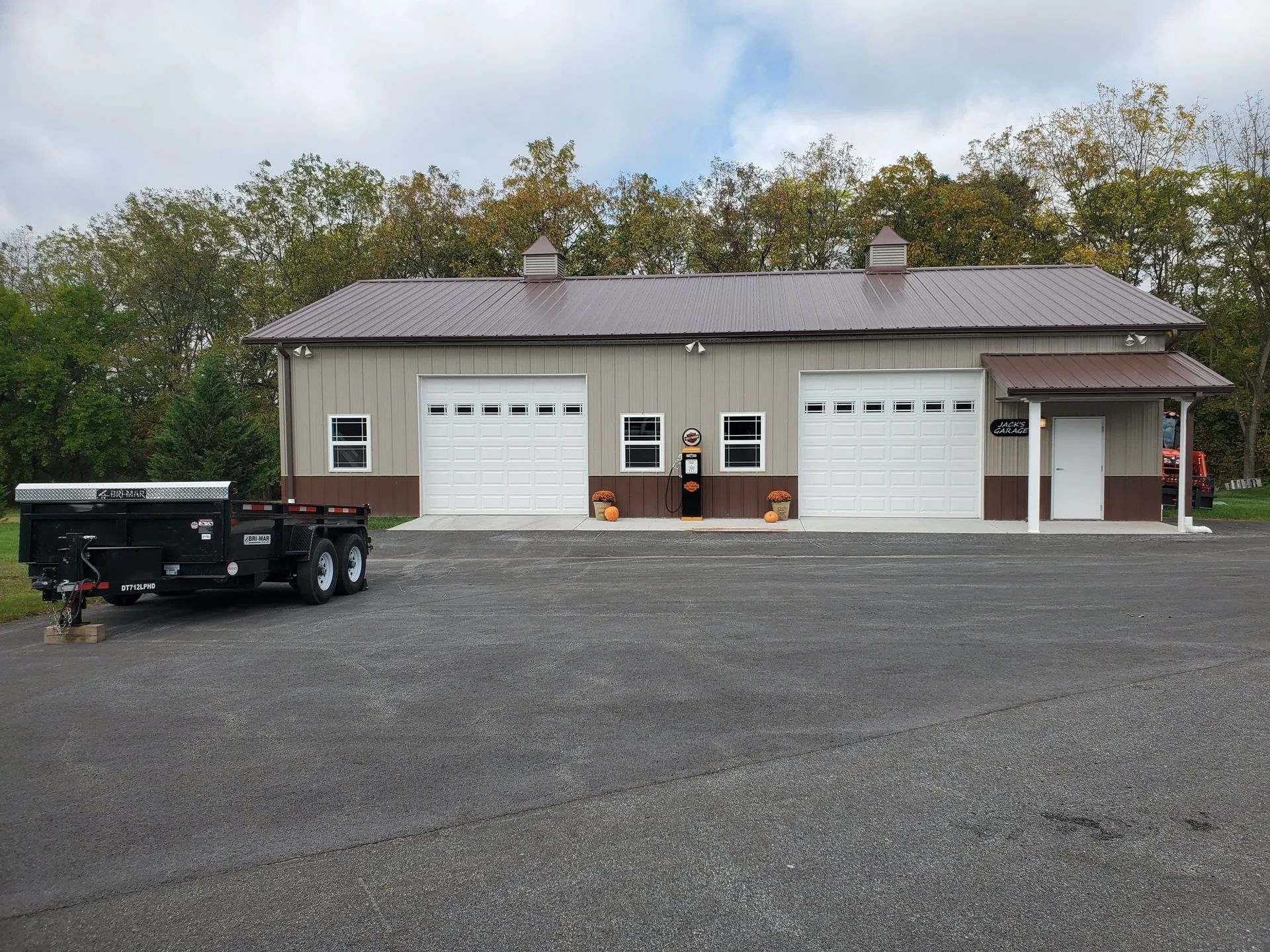 A wide-angle shot of a tan, two-car garage building with a brown metal roof, set against a backdrop of trees.