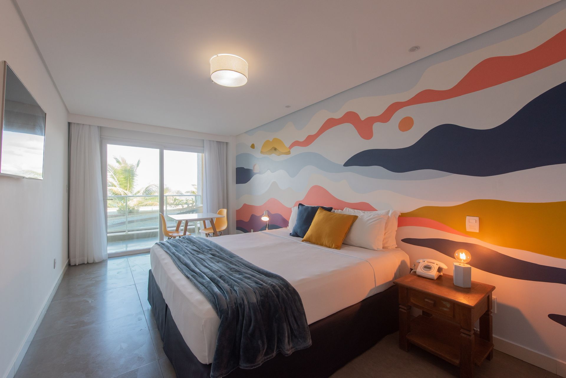 Quarto aconchegante com cama de casal, decoração moderna, varanda e vista para coqueiros em ambiente iluminado.