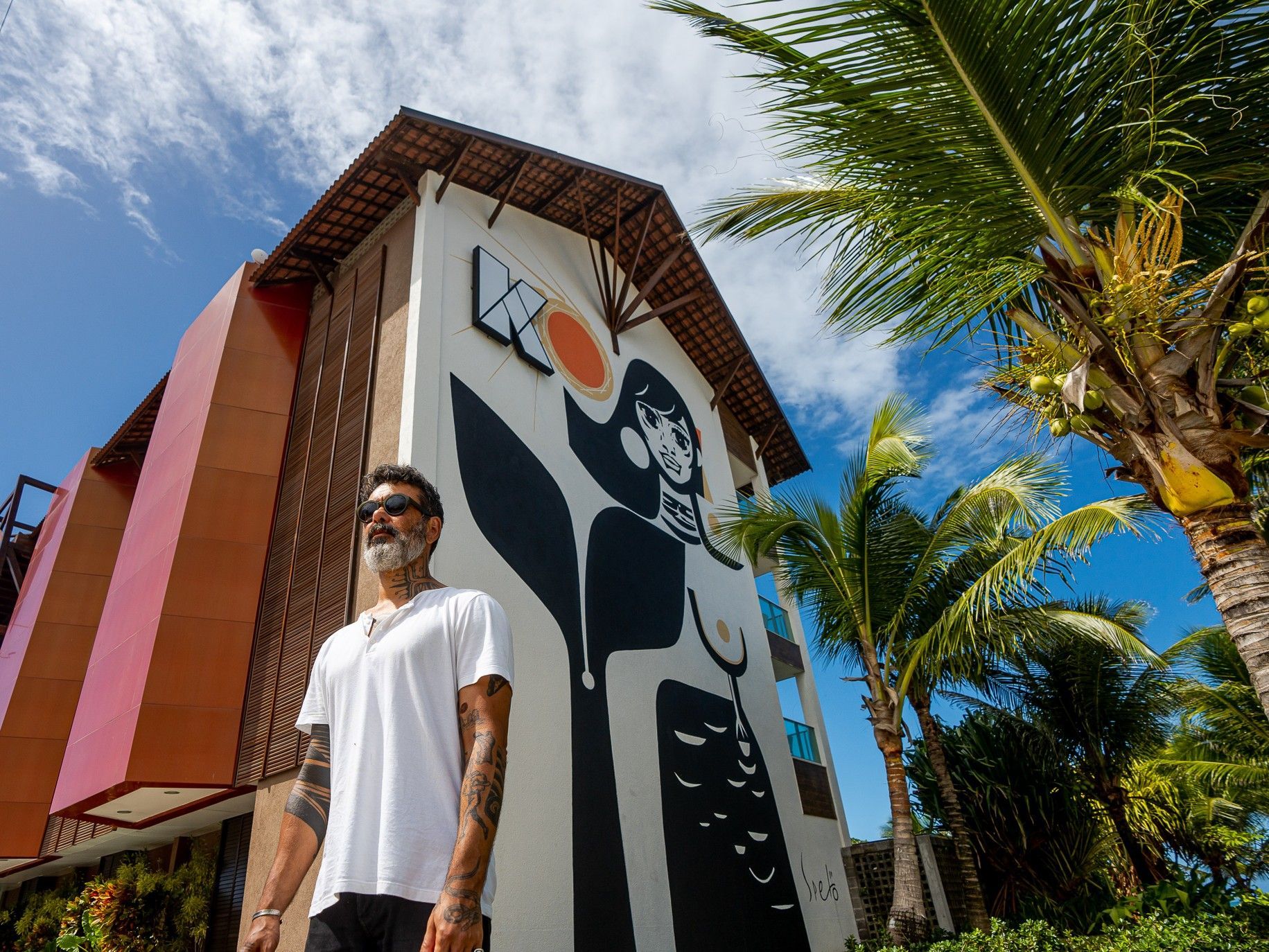 Um artista barbudo com óculos escuros e camiseta branca posa em frente a um prédio. Na fachada, há um grande mural em preto e branco com detalhes em laranja, retratando uma figura feminina estilizada.