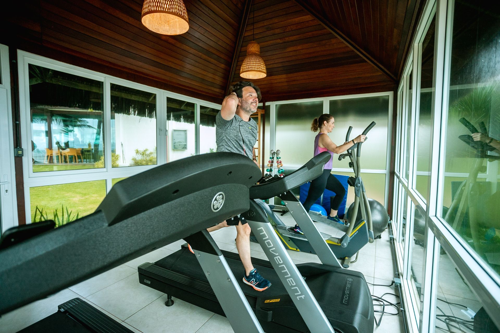 A imagem mostra casal em academia do hotel. O homem caminha na esteira e a mulher utiliza a máquina de elíptico. O teto é de madeira com luminárias em palha e as paredes são de vidro, mostrando o exterior com gramado.
