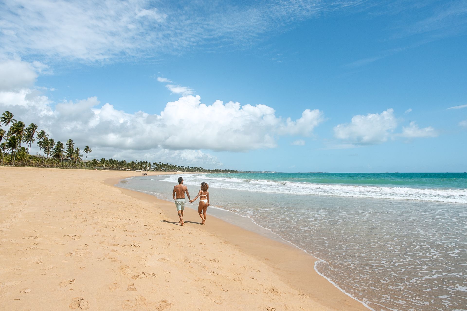 Homem e mulher caminhando na areia da praia. O mar está azul, o céu está azul e com algumas nuvens brancas. Ao fundo é possível ver coqueiros.