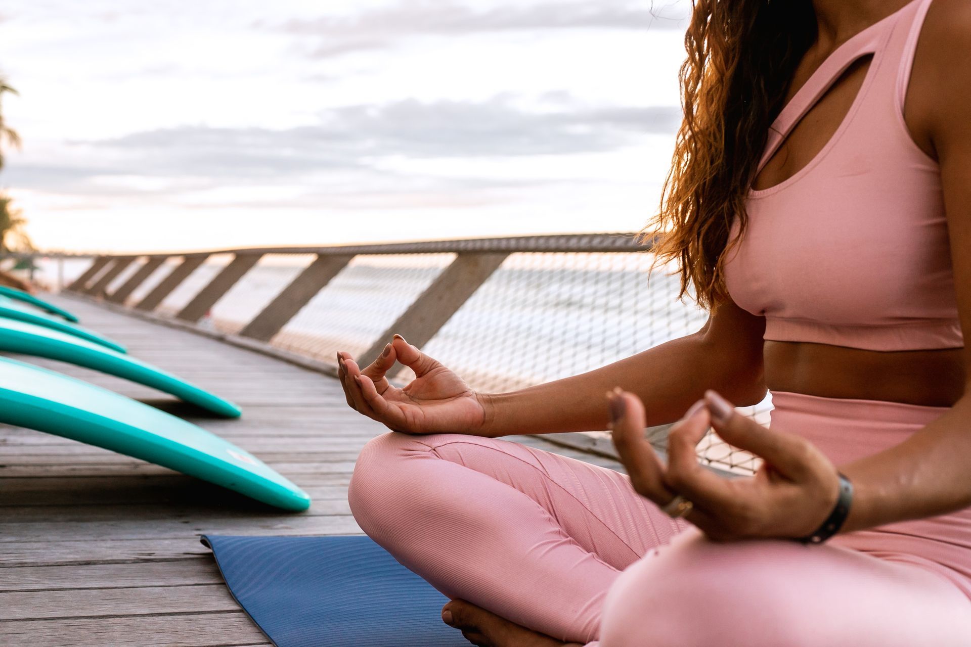 Foto mostra mulher em posição de meditação de yoga. Ela está sentada no deck de madeira do hotel, com o mar ao fundo, em fim de tarde. Ela está com as pernas cruzadas e as mãos apoiadas no joelho, com o dedo indicador e polegar conectados. Ela usa legging e top na cor rosa claro.