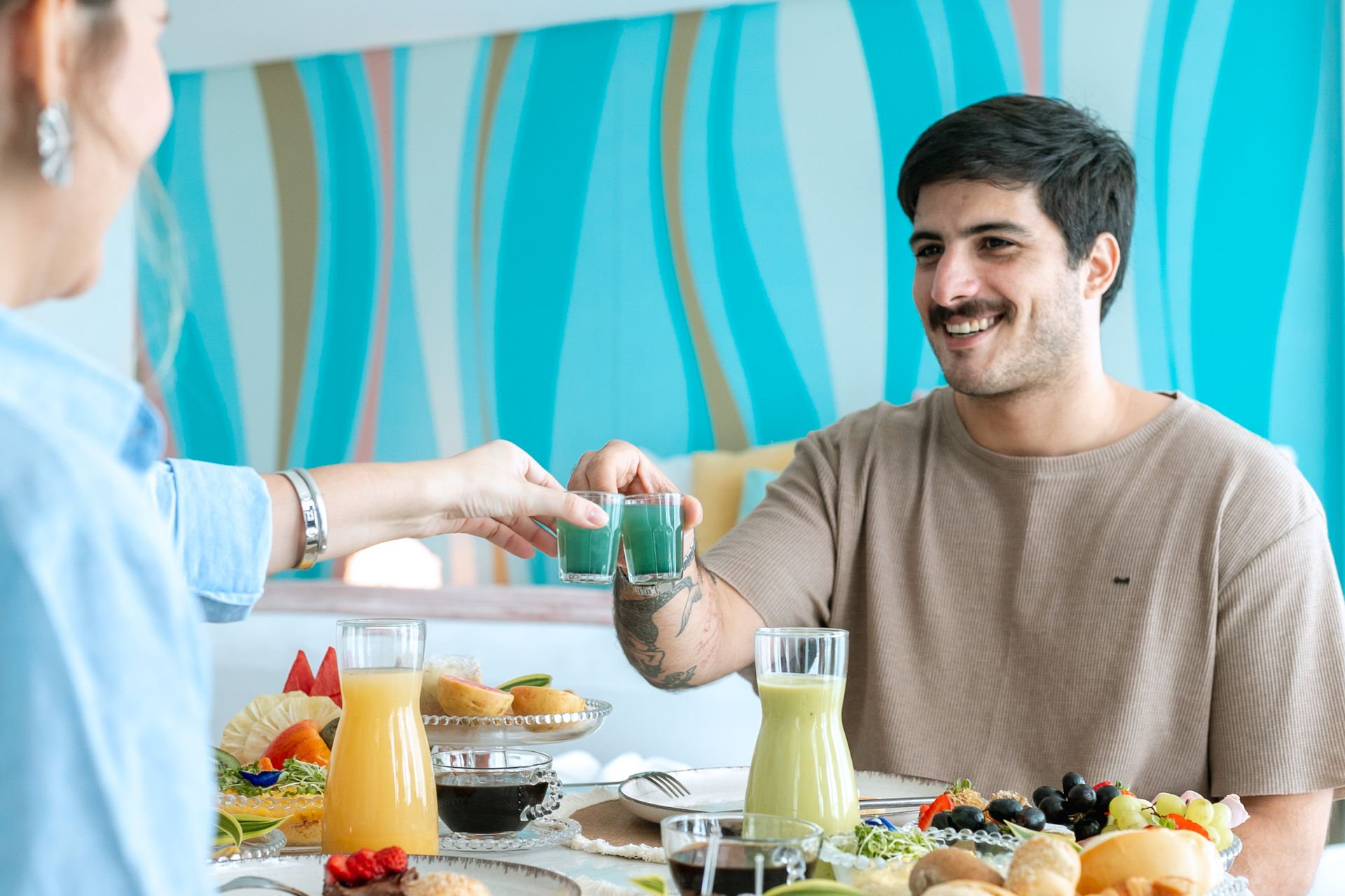 Casal tomando café da manhã no quarto do hotel e os dois estão brindando com shot da imunidade na cor azul. A mesa do café da manhã esá cheia de frutas, pães e sucos.
