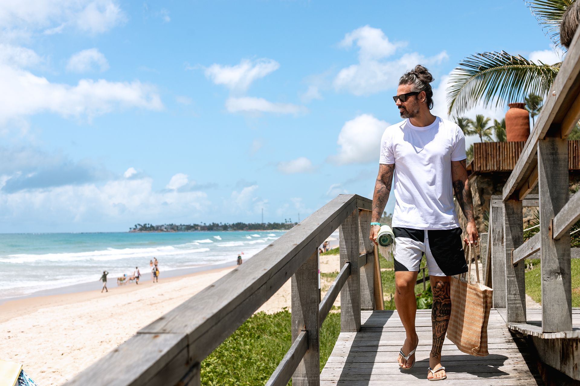 Homem caminhando pelo acesso à praia com bolsa e toalha, em cenário à beira-mar de Porto de Galinhas.
